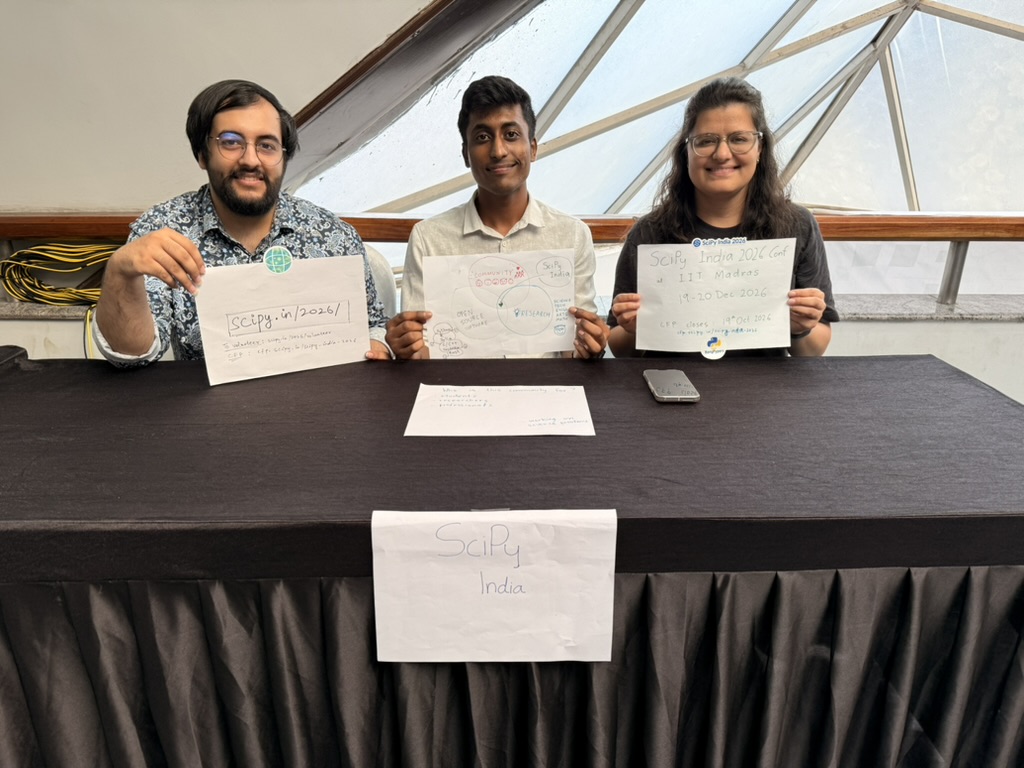 Agriya, Srihari, and Malayaja sitting at the SciPy India booth holding up handwritten signs