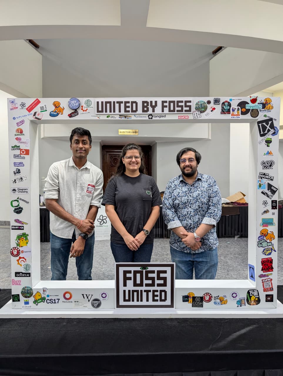 Srihari, Malayaja, and Agriya standing inside the sticker-covered United by FOSS photo frame